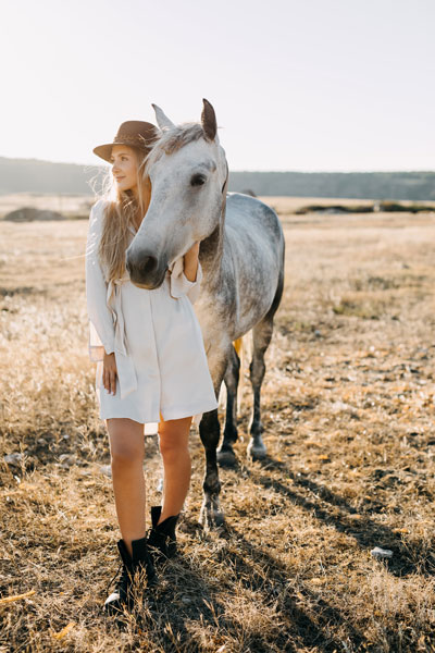 Young pregnant woman cowgirl with white horse standing in a field. 